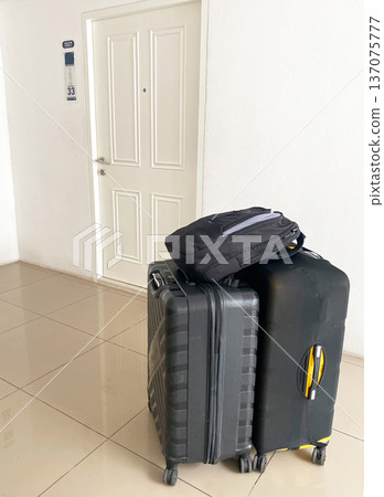 Two pieces of luggage stacked beside a closed white door in a rented apartment hallway, with a black backpack resting on top of a hard-shell suitcase 137075777