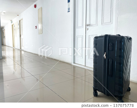 Black suitcase positioned in a well-lit hotel corridor with white walls, numbered door visible, and polished tile floor reflecting light 137075778