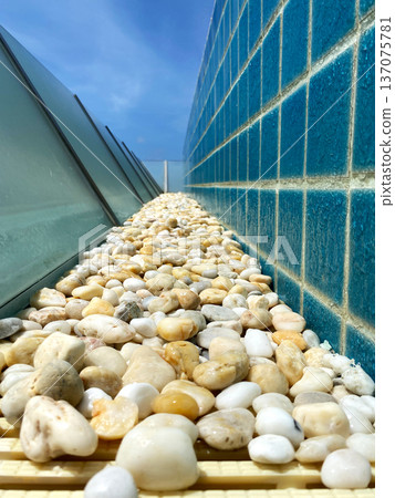 Decorative gravel pathway lined with smooth pebbles alongside a blue tiled wall, featuring a clear sky and modern architectural elements in a pool area 137075781