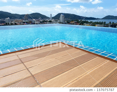 Infinity pool with wooden deck overlooking cityscape and mountains under a clear blue sky, featuring calm water and vibrant urban architecture in the background 137075787