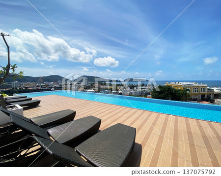 Infinity pool with lounge chairs overlooking cityscape and ocean under blue sky with clouds, tropical landscape in the background, Phuket, Thailand Infinity pool with lounge chairs overlooking cityscape and ocean under blue sky with clouds, tropical landscape in the background, Phuket, Thailand 137075792