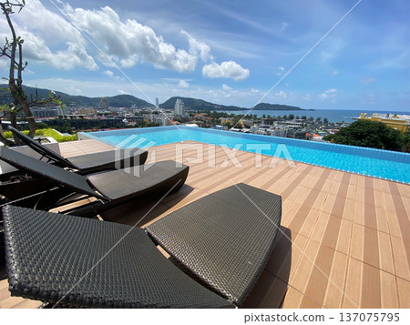 Luxury outdoor poolside view with modern lounge chairs overlooking the ocean and cityscape, surrounded by lush greenery and mountains in the background 137075795