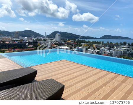 Rooftop swimming pool with infinity edge overlooking cityscape and mountains, featuring wooden deck and lounge chairs under a clear blue sky Rooftop swimming pool with infinity edge overlooking cityscape and mountains, featuring wooden deck and lounge chairs under a clear blue sky 137075796