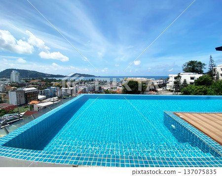 Rooftop swimming pool with clear blue water overlooking a coastal cityscape, featuring buildings, trees, and distant mountains under a bright sky Rooftop swimming pool with clear blue water overlooking a coastal cityscape, featuring buildings, trees, and distant mountains under a bright sky 137075835