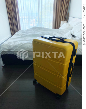 Yellow suitcase placed beside an unmade bed in a hotel room with large windows allowing natural light to illuminate the space 137075845