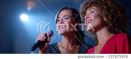 Two young women singing together on stage with microphone under bright spotlight in a joyful performance setting 137075976