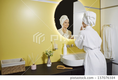 Happy beautiful woman looking in modern bathroom mirror, wearing white robe and towel wrapped 137076384