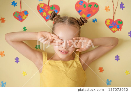 A shy, modest girl with autism covers her face with her hands. The background is decorated with hearts and pieces of colorful colorful puzzles - an autistic symbol A shy, modest girl with autism covers her face with her hands. The background is decorated with hearts and pieces of colorful colorful puzzles - an autistic symbol 137078950