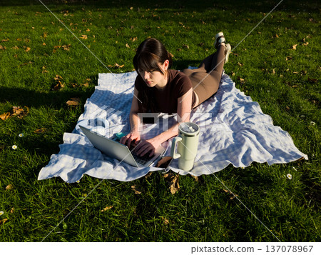 Young woman working on laptop outdoors in park, remote freelance lifestyle, female typing on computer while lying on blanket, digital nomad productivity concept 137078967