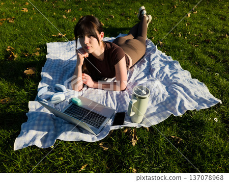 Young woman working remotely on laptop in sunny park, freelance digital nomad lifestyle, female typing on computer outdoors on blanket in green grass Young woman working remotely on laptop in sunny park, freelance digital nomad lifestyle, female typing on computer outdoors on blanket in green grass 137078968