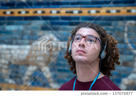 Berlin, germany, august 16, 2023. Young man with headphones and glasses gazes up at the blue-tiled ishtar gate in the pergamon museum, absorbed in history and culture 137078977