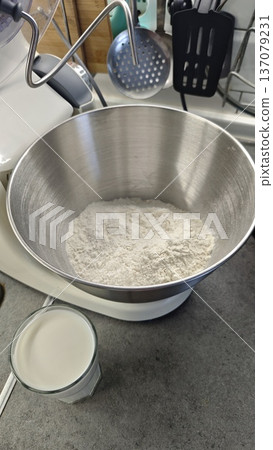 Flour in metal mixer bowl with dough hook and glass of milk on kitchen countertop. Baking preparation and homemade bread recipe concept Flour in metal mixer bowl with dough hook and glass of milk on kitchen countertop. Baking preparation and homemade bread recipe concept 137079231