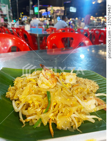Plate of stir-fried noodles with shrimp, bean sprouts, and vegetables served on a banana leaf at a vibrant outdoor food market with red plastic chairs Plate of stir-fried noodles with shrimp, bean sprouts, and vegetables served on a banana leaf at a vibrant outdoor food market with red plastic chairs 137079467