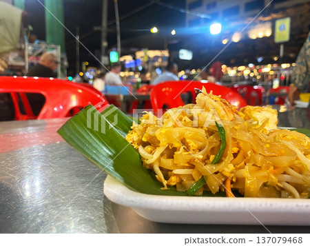Pad Thai served on a white plate with banana leaf garnish at a bustling night market restaurant with red plastic chairs and vibrant lights in the background Pad Thai served on a white plate with banana leaf garnish at a bustling night market restaurant with red plastic chairs and vibrant lights in the background 137079468