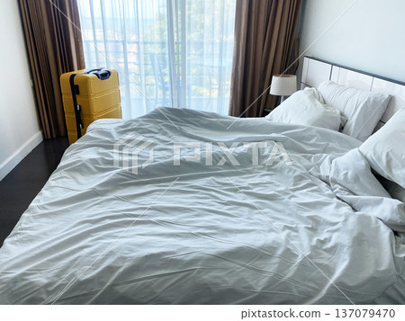Unmade bed with crumpled white sheets and yellow suitcase in a hotel room, large window with sheer curtains allowing natural light to enter Unmade bed with crumpled white sheets and yellow suitcase in a hotel room, large window with sheer curtains allowing natural light to enter 137079470