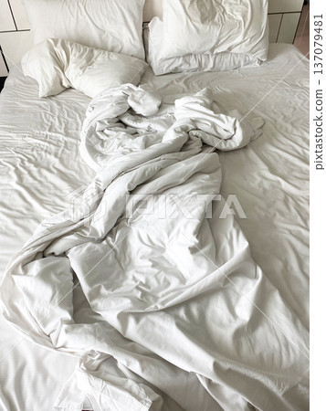 Unmade wrinkled white bed linen with crumpled sheets and pillows on a bed, showcasing a casual and relaxed bedroom setting 137079481