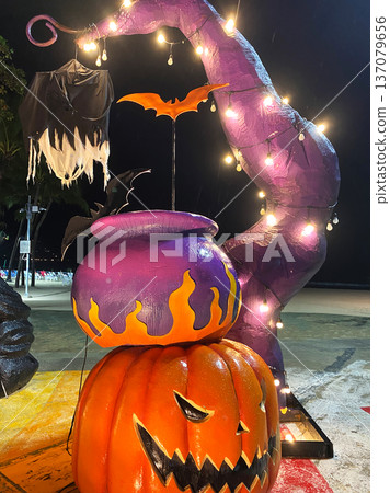 Halloween illuminated decorations featuring a large orange pumpkin, purple cauldron, and bat motifs in a city center at night with string lights glowing Halloween illuminated decorations featuring a large orange pumpkin, purple cauldron, and bat motifs in a city center at night with string lights glowing 137079656