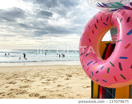 Pink donut-shaped inflatable float placed on sandy beach with people swimming in the ocean under a cloudy sky during daytime 137079675