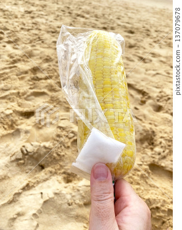 Boiled corn on the beach held in a hand, wrapped in plastic with salt packet, sandy background and ocean waves visible in the distance 137079678