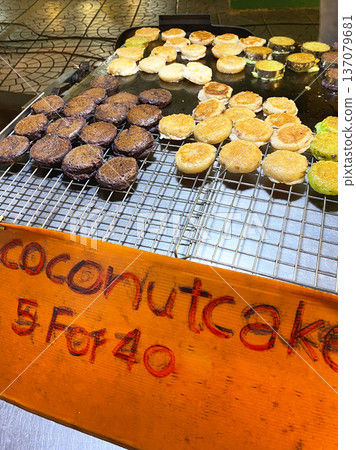 Coconut pancakes displayed on a wire rack at a street food stall in Asia, with a colorful sign indicating pricing and variety of flavors visible Coconut pancakes displayed on a wire rack at a street food stall in Asia, with a colorful sign indicating pricing and variety of flavors visible 137079681