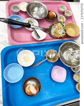 Dirty dishes piled on pink and blue trays after cooking, featuring various bowls, utensils, and remnants of food, creating a cluttered kitchen scene Dirty dishes piled on pink and blue trays after cooking, featuring various bowls, utensils, and remnants of food, creating a cluttered kitchen scene 137079770