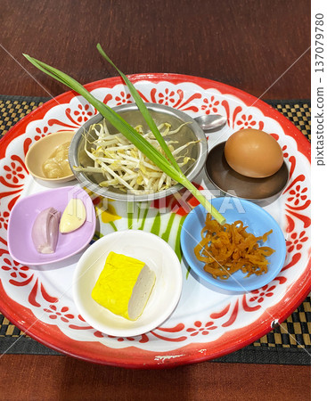 Ingredients for Pad Thai arranged on a decorative plate, including bean sprouts, egg, tofu, garlic, and green onions, with colorful bowls and utensils visible Ingredients for Pad Thai arranged on a decorative plate, including bean sprouts, egg, tofu, garlic, and green onions, with colorful bowls and utensils visible 137079780