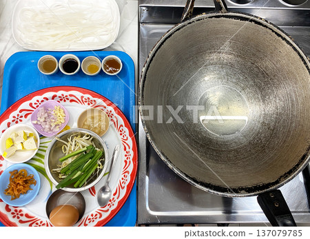 Ingredients for making classic Pad Thai arranged on a decorative plate with a large wok and cooking utensils visible in a kitchen setting 137079785