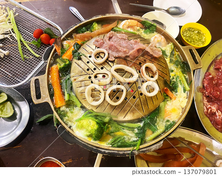 Hot pot restaurant scene featuring a metal pot filled with broth, vegetables, seafood, and meat, surrounded by various ingredients on a wooden table 137079801