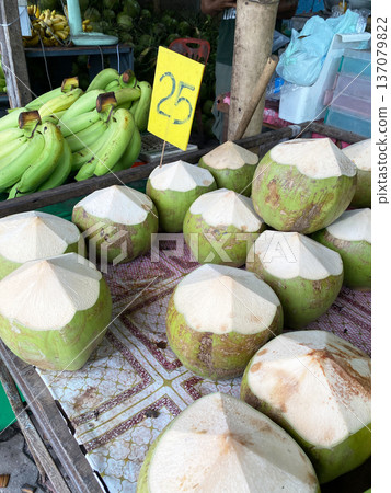 Fresh green coconuts with cut tops displayed on a market table, with a price sign and bunch of bananas in the background under natural lighting Fresh green coconuts with cut tops displayed on a market table, with a price sign and bunch of bananas in the background under natural lighting 137079822