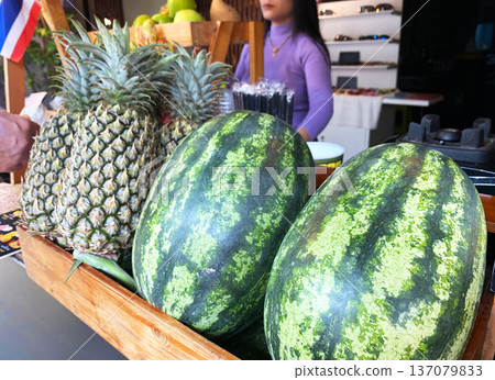 Freshly harvested watermelons and pineapples displayed in a wooden crate at a juice stand with a woman preparing drinks in the background Freshly harvested watermelons and pineapples displayed in a wooden crate at a juice stand with a woman preparing drinks in the background 137079833