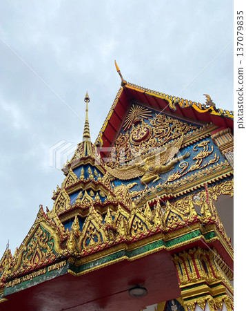 Ornate temple structure in Thailand featuring intricate gold decorations, colorful murals, and traditional architectural elements against a cloudy sky backdrop Ornate temple structure in Thailand featuring intricate gold decorations, colorful murals, and traditional architectural elements against a cloudy sky backdrop 137079835