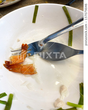 Empty white plate with leftover shrimp, fork, and spoon, garnished with green onion pieces, showcasing remnants of a meal in a casual dining setting 137079846
