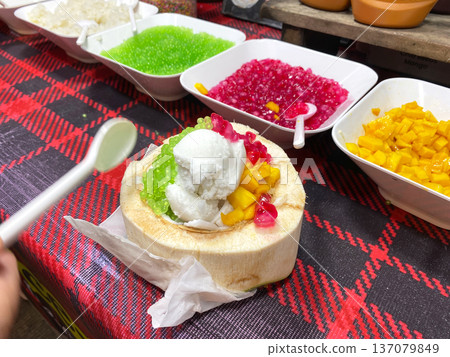 Coconut ice cream served in a halved coconut shell topped with colorful fruit and jelly, surrounded by bowls of additional toppings on a checkered tablecloth Coconut ice cream served in a halved coconut shell topped with colorful fruit and jelly, surrounded by bowls of additional toppings on a checkered tablecloth 137079849