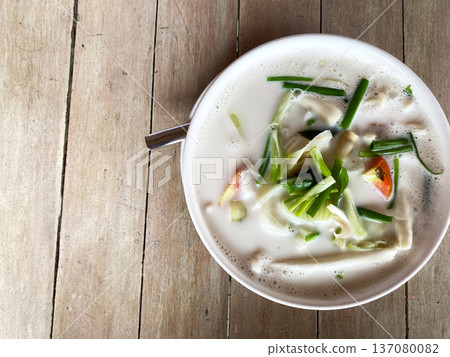 Coconut tom yum soup served in a white bowl with green onions and vegetables on a rustic wooden table background, showcasing vibrant colors and textures Coconut tom yum soup served in a white bowl with green onions and vegetables on a rustic wooden table background, showcasing vibrant colors and textures 137080082