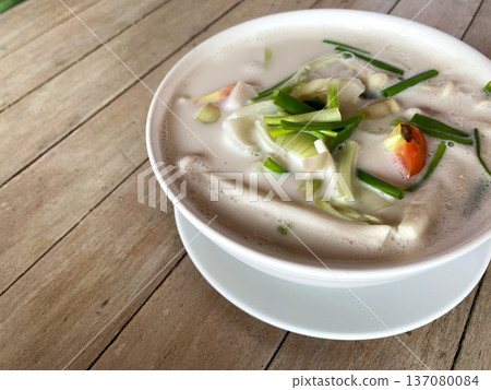 Coconut tom yum soup in a white bowl garnished with green onions and vegetables on a rustic wooden table background, showcasing vibrant colors and textures Coconut tom yum soup in a white bowl garnished with green onions and vegetables on a rustic wooden table background, showcasing vibrant colors and textures 137080084