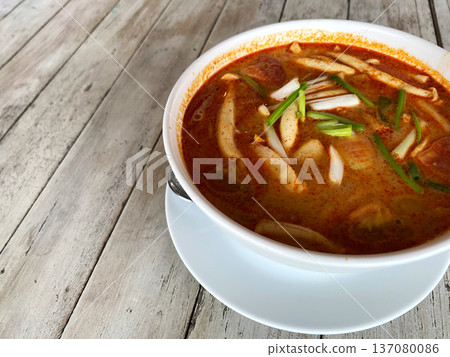 Tom Yum soup served in a white bowl with green onions on a wooden table, showcasing vibrant colors and rich textures of the dish and background Tom Yum soup served in a white bowl with green onions on a wooden table, showcasing vibrant colors and rich textures of the dish and background 137080086