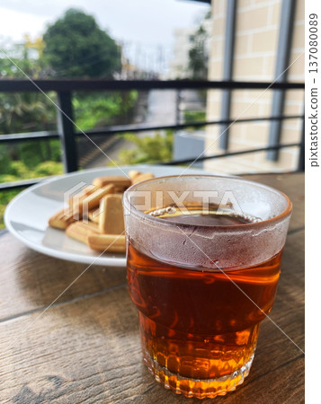 Glass of amber tea placed on wooden table beside a plate of assorted cookies, with greenery visible through a balcony railing in the background Glass of amber tea placed on wooden table beside a plate of assorted cookies, with greenery visible through a balcony railing in the background 137080089