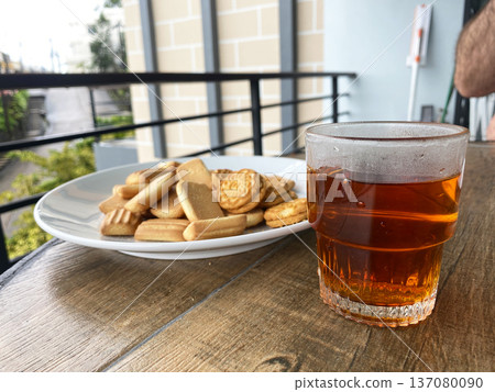 Glass of amber tea beside a plate of assorted cookies on a wooden table with a blurred outdoor background featuring greenery and a railing Glass of amber tea beside a plate of assorted cookies on a wooden table with a blurred outdoor background featuring greenery and a railing 137080090