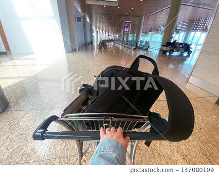 Woman's hand grips a luggage cart with a black backpack inside an airport terminal, bright lighting and modern architecture visible in the background 137080109