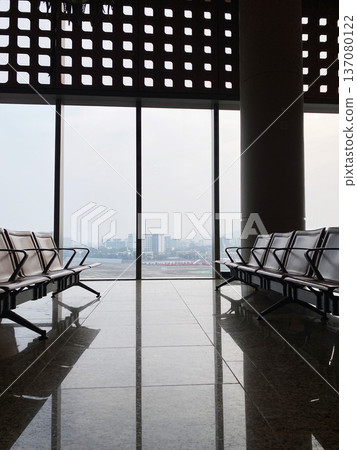 Modern airport terminal interior featuring empty seating rows with large glass windows showcasing an urban skyline in the background and polished marble flooring 137080122