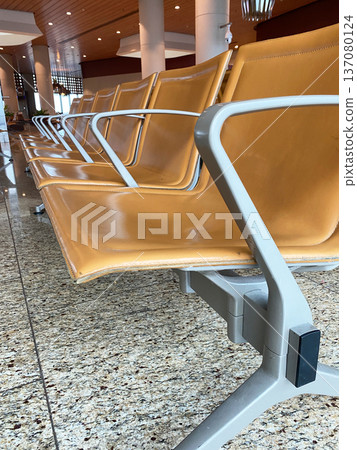 Airport waiting area featuring modern orange seating with metal frames, polished granite flooring, and bright overhead lighting in a spacious terminal environment Airport waiting area featuring modern orange seating with metal frames, polished granite flooring, and bright overhead lighting in a spacious terminal environment 137080124