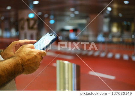 Person using smartphone to call a taxi at an airport terminal with red flooring and blurred background showing vehicle pick-up area 137080149