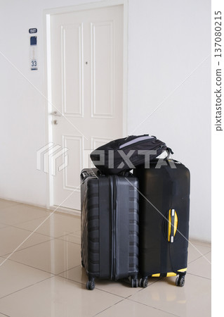 Two large suitcases and a black backpack stacked near a white door with a room number sign, indicating a temporary stay in an apartment or hotel Two large suitcases and a black backpack stacked near a white door with a room number sign, indicating a temporary stay in an apartment or hotel 137080215