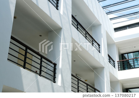 Modern architectural design featuring white walls and balconies with black railings in a well-lit interior space with large windows and natural light Modern architectural design featuring white walls and balconies with black railings in a well-lit interior space with large windows and natural light 137080217