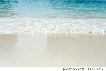 Calm turquoise ocean waves gently lapping onto a pristine sandy beach under bright sunlight, creating a serene coastal landscape with clear blue skies 137080247