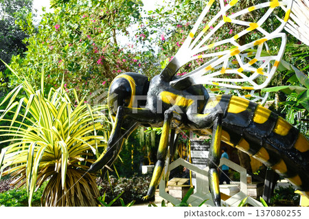 Close-up model of a bee sculpture with yellow and black stripes, surrounded by tropical plants and flowers in a vibrant garden setting 137080255
