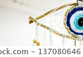Crocheted evil eye dreamcatcher featuring a detailed blue and white eye pattern within a woven wooden frame and hanging white ribbons with wooden beads. Symbolizing protection and spiritual well being 137080646