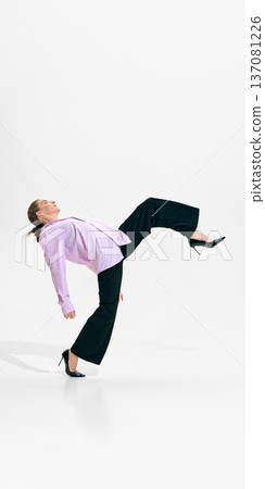Woman in lavender shirt and black trousers balancing on one leg on white studio background 137081226