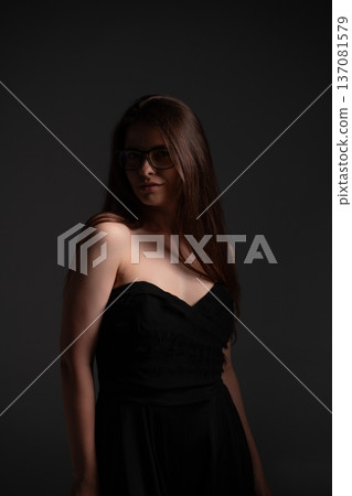Stylish woman wearing glasses and black strapless dress posing with long brown hair, creating mysterious and elegant low key studio portrait for fashion and beauty concepts Stylish woman wearing glasses and black strapless dress posing with long brown hair, creating mysterious and elegant low key studio portrait for fashion and beauty concepts 137081579