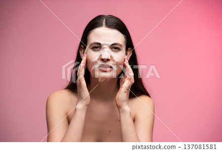 Woman applying a refreshing sheet face mask for clear, glowing skin, focusing on beauty, self-care, and a healthy skincare routine against a pink background 137081581